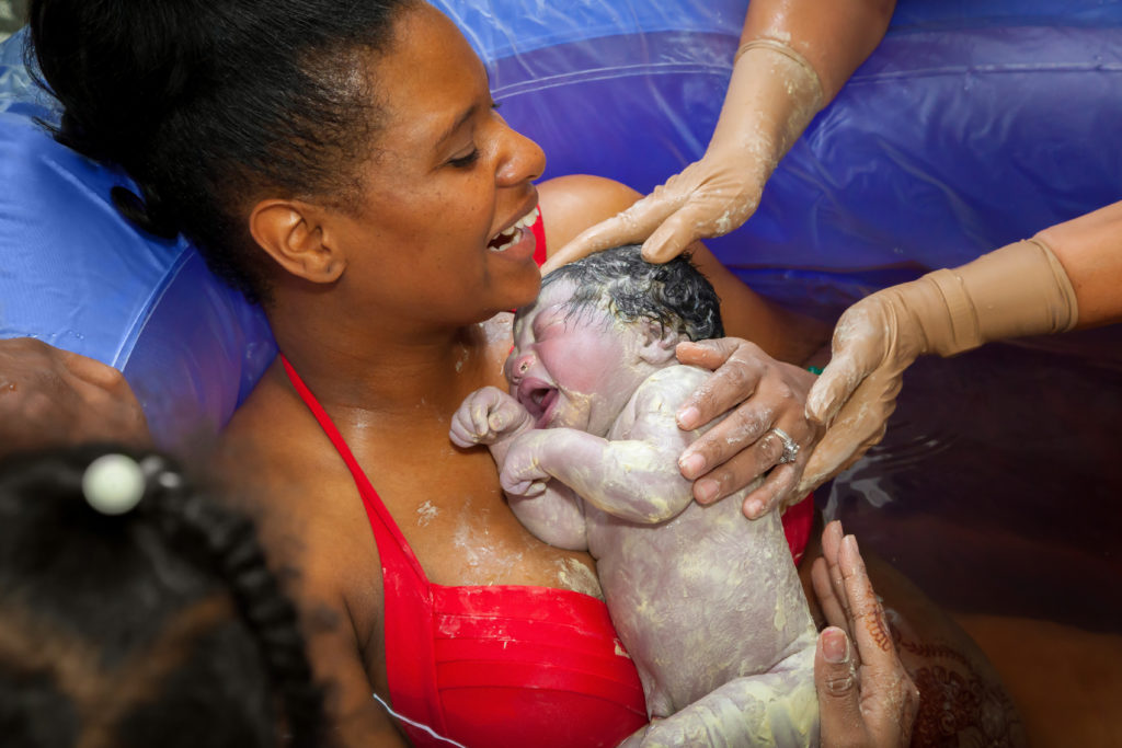 A happy African American mother holding her brand new baby girl after delivering her in a birthing pool at home. The baby is covered in vernix and the mom is laughing.